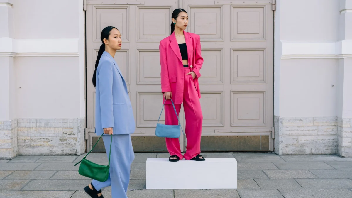 Twee modieuze vrouwen in levendige blazers poseren met stijlvolle handtassen voor een vintage gebouw.