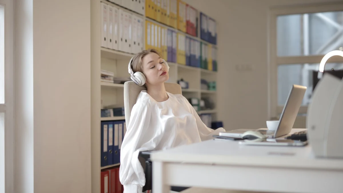 Vrouw in witte overhemd ontspannend aan kantoor bureau met koptelefoon, lijkt rustgevend.